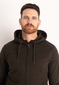 Felpa con zip di un verde scuro, dotata di cappuccio regolabile con coulisse, polsini a costine e texture morbida. Presenta una chiusura con zip e un design minimale.