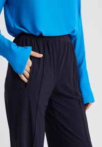 Nahaufnahme einer Person, die schwarze Hosen mit elastischem Bund und ein hellblaues Langarmshirt trägt, die Hand ruht in der Hosentasche.