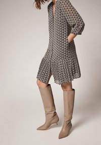 Robe à motif géométrique en noir et beige, jupe plissée, manches trois-quarts. Associée à des bottes beige pointues montant jusqu'aux genoux.