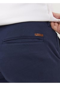 Närbild av en marinblå byxficka med en liten brun läderlogga och en vit skjorta delvis instoppad.