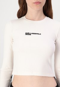 Bijela rebrasta dugorukava crop majica s crnim vezom loga "KARL LAGERFELD JEANS" i bočnim detaljima s brendingom.