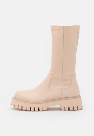 Botas de goma beige hasta la rodilla con un acabado texturizado, que cuentan con una suela gruesa y paneles laterales elásticos para facilitar su uso. Diseño minimalista.
