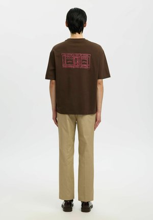Homme aux cheveux noirs vu de dos, portant un t-shirt marron avec un motif de devanture rouge et un pantalon beige, sur fond blanc uni.