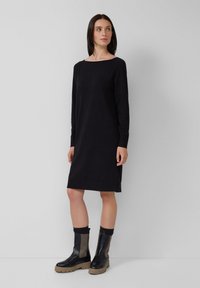 Vestido negro de manga larga con escote barco, ajuste ligeramente holgado, con aberturas laterales, combinado con botas de tobillo gruesas en negro y gris.