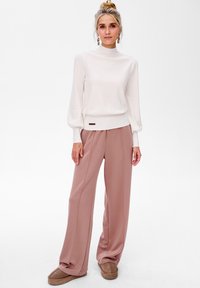 Lekki, biały sweter z wysokim dekoltem, długimi rękawami i ściągaczem; zestawiony z luźnymi, miękkimi różowymi spodniami i brązowymi klapkami.