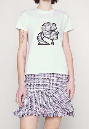 Frau trägt ein weißes T-Shirt mit einer Doppel-Silhouetten-Grafik kombiniert mit einem mehrfarbigen Tweed-Minirock mit Rüschensaum.