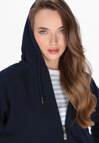 Námořnická mikina na zip s kapucí na šňůrku, se stříbrným zipem a pod ní pruhovaná bílá a modrá košile. Viditelné dlouhé vlnité vlasy.
