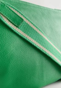 Grüne Leder-Tote mit einer strukturierten Oberfläche, ausgestattet mit einem kontrastierenden beigen gestreifen Stoffgurt. Reißverschluss an der oberen Kante.