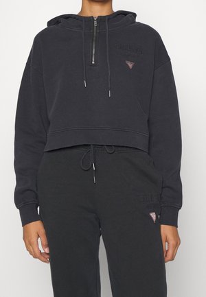 Personne portant un sweat à capuche court noir avec fermeture éclair à l'avant et un pantalon de jogging noir assorti, avec des logos Guess discrets et des cordons.