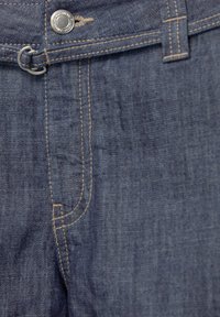 Denimtyg i mörkblått, med kontrasterande ljusa sömmar och en knappstängning med ögla. Närbild på midjeområdet.