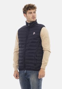 Gilet imbottito blu navy con colletto alto, chiusura con zip e cuciture orizzontali. Indossato sopra un maglione beige, presenta un logo sul petto.
