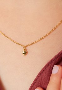 Gouden halsketting met een delicate ketting en een kleine, glanzende hartvormige hanger. De achtergrond heeft een zachte huidtint en stoffen textuur.