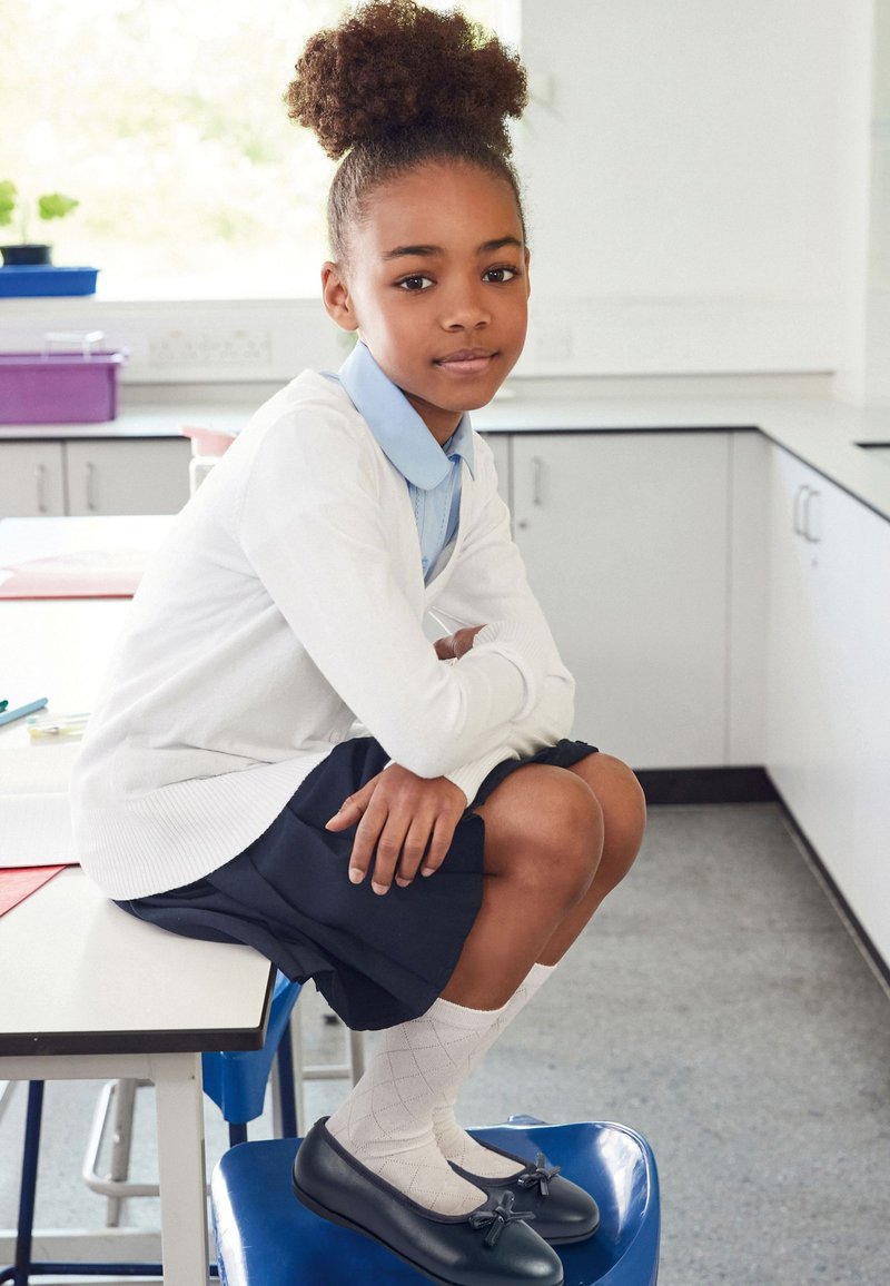 Schwarze Lacklederschuhe mit Schleifendetail, weißer Cardigan, blaues Hemd und dunkelblauer Rock. Das Mädchen sitzt auf einem blauen Hocker in einem Klassenzimmer.