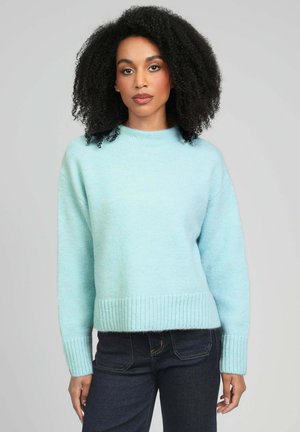 Femme aux cheveux bouclés portant un pull bleu clair et un jean foncé, debout devant un fond gris clair uni.