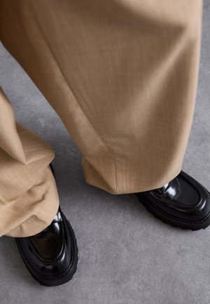 Beige bukser med en tekstureret overflade, parret med blanke sorte loafers med en tyk sål og kontrastsyning.