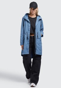 Jachetă lungă de un bleu deschis cu glugă, fermoar frontal și şireturi. Pur și simplu peste un top negru scurt și pantaloni cargo negri, asortați cu adidași albi.