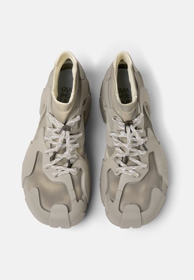 Sneakers grigi chiaro con una tomaia in rete testurizzata, sovrastrutture in gomma e lacci bianchi piatti. Presentano un design unico e massiccio con ritagli distintivi.