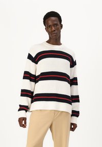 Suéter a rayas en crema, marino y rojo. Textura acanalada, escote redondo y diseño de hombros caídos. Combinado con pantalones beige.