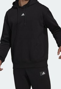 Persona che indossa un'hoodie nera Adidas e pantaloni neri abbinati con i loghi Adidas sul petto e sulla coscia.