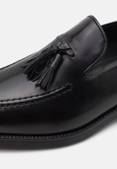 Mocassins en cuir noir avec un accent à franges et des détails cousus sur le dessus, à la texture lisse et avec une semelle plate en caoutchouc.