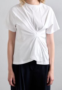 Bílý bavlněný tričko s propleteným designem na přední straně, krátké rukávy a uvolněný střih, kombinované s tmavými kalhotami.