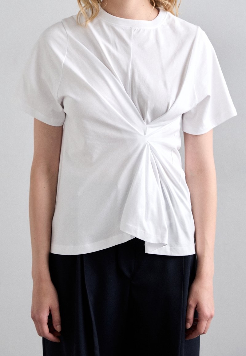 Bílý bavlněný tričko s propleteným designem na přední straně, krátké rukávy a uvolněný střih, kombinované s tmavými kalhotami.