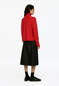 Eine Frau mit langen dunklen Haaren steht seitlich und trägt einen roten Rollkragenpullover, einen schwarzen Midikleid, schwarze Socken und schwarze glänzende Schuhe.