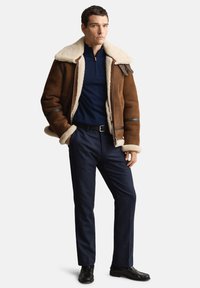 Bruine shearling jas met crèmekleurige voering, marineblauwe pullover, donkere broek en zwarte schoenen. Voorzien van rits en leren accenten.