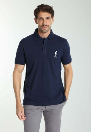 Homme portant un polo bleu marine avec un logo de cheval blanc et un pantalon gris clair, debout sur un fond uni clair.