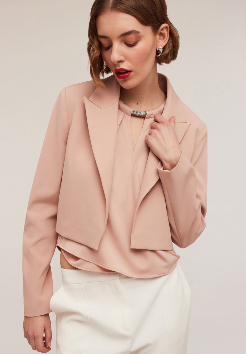 Blazer corti rosa pallido con rever a punta, maniche lunghe e tessuto liscio, abbinato a una blusa coordinata e pantaloni bianchi a vita alta.