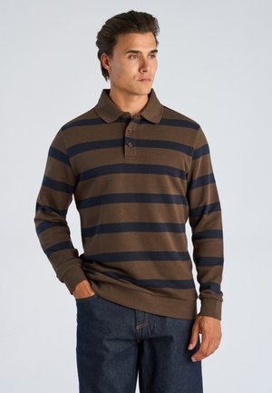 Mand iført brun og marineblå stribet langærmet poloshirt med tre knapper, stående med den ene hånd i lommen og kiggende mod højre.