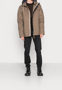 Hnědá voděodolná bunda s kapucí, zapínání na zip, černé pruhované tričko pod ní, černé kalhoty a černé šněrovací boty. Jednoduchý design.