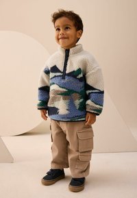 Ungt barn iført en mønstret fleecepullover, beige cargo-bukser og sorte sneakers, stående i et neutralt indendørsmiljø.