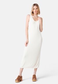 Knit white maxi dress with a scoop neckline, form-fitting shape, ribbed texture, and side slit. Paired with brown heeled clogs.