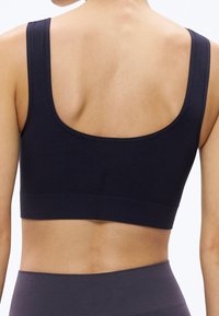 Moteris, vilkinti juodą sportinį liemenėlę ir tamsiai pilkus aukštos juosmens sportinius leginsus, nugaros vaizdas prieš baltą foną.