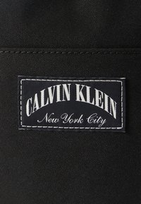 Μαύρη ετικέτα από ύφασμα με λευκό κείμενο «CALVIN KLEIN» σε τόξο και «New York City» σε μικρότερη, καλλιγραφική γραμματοσειρά, περιτριγυρισμένη από λευκή ραφή.