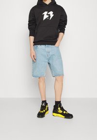 Felpa nera con logo a fulmine bianco, pantaloni shorts in denim leggero blu, e sneakers nere con dettagli gialli, esposti su uno sfondo semplice.