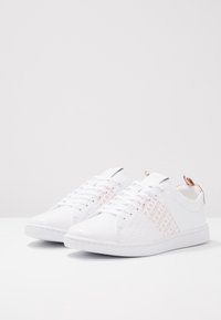 Para białych, niskich trampków z białymi sznurowadłami i wzorem tekstowym w kolorze różowego złota "Lacoste Paris" po bokach, na jednolitym tle.