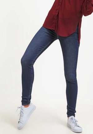 Person trägt dunkelblaue Skinny Jeans, weiße Sneakers und ein bordeauxfarbenes Hemd mit Knöpfen und seitlichen Schlitzen und posiert mit einem angewinkelten Bein.