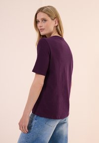T-shirt viola a maniche corte in un tessuto morbido, con collo rotondo e vestibilità rilassata, abbinata a jeans in denim azzurri chiari.