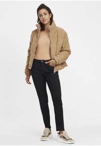 Veste en velours côtelé tan avec un col haut, pull léger marron clair à côtes, jeans slim noirs et baskets à bord beige. Look simple et décontracté.