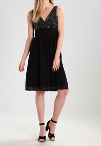 Svart ärmlös klänning med V-ringad hals, utsmyckad överdel och flödande kjol. Har ett brett midjeband och genomskinlig nederkant. Bärs med svarta klackar.