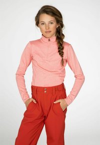 Langsleeve, roze top met rits, gemaakt van textuurstof, gecombineerd met hoge taille, rode broek met zijzakken en metalen sluitingen.