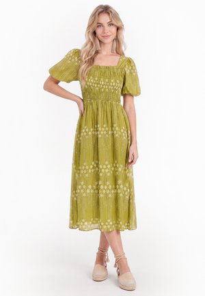 Kvinde stående med hånd på hoften iført en grøn blomster-midi kjole med pufærmer og beige espadrillosko med ankelsnører.