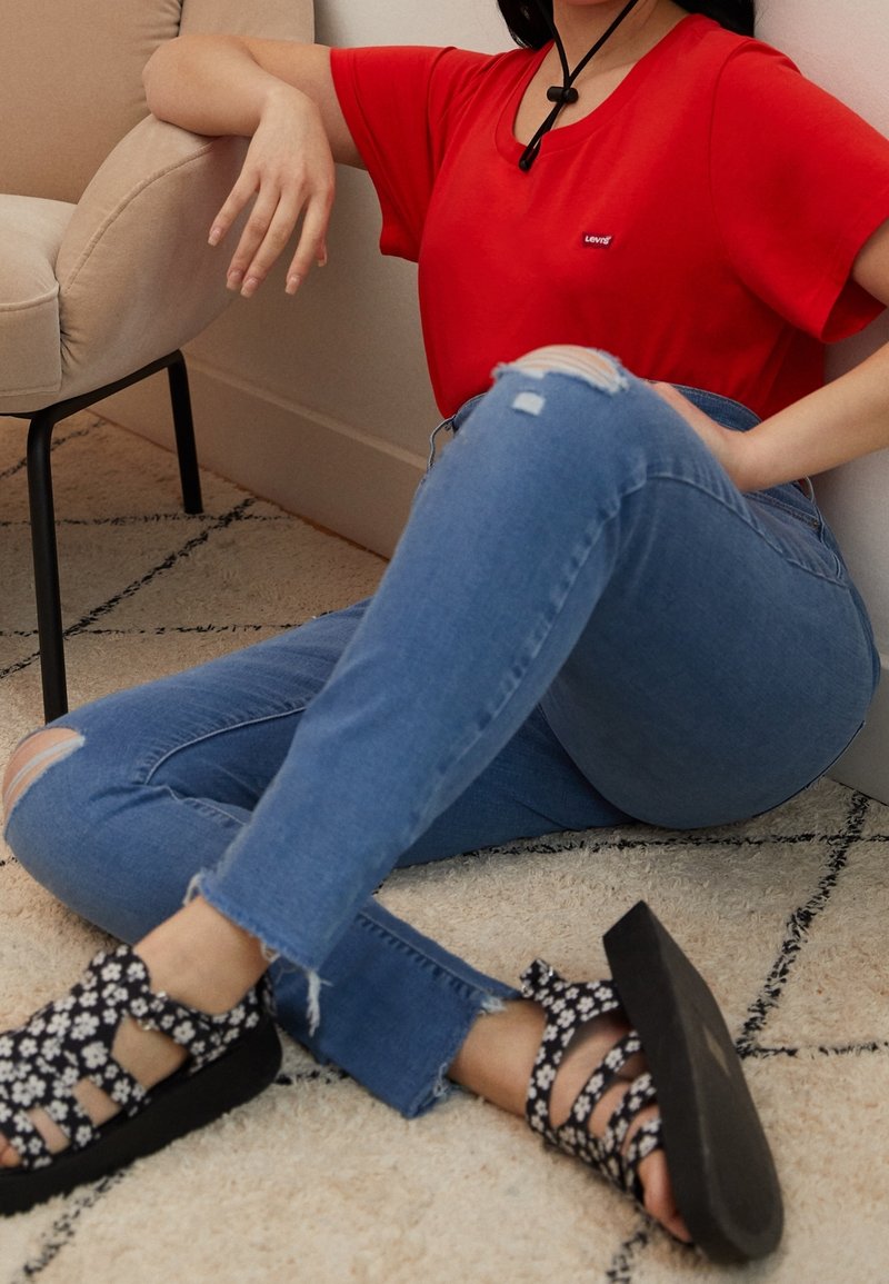 T-shirt rouge à manches courtes, jeans déchirés bleu clair et sandales noires à fleurs. Position assise sur un tapis crème texturé avec un fauteuil de couleur claire.