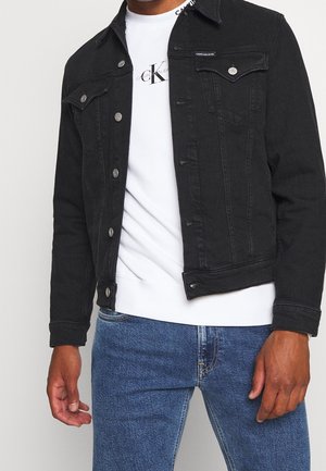 Muž oblečený v černé džínové bundě přes bílou mikinu Calvin Klein a modré džíny na jednobarevném pozadí.