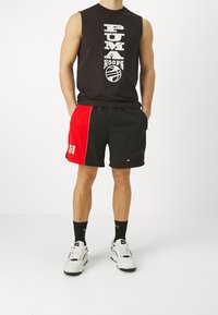 Muž oblečený v černém bezrukávovém tričku s nápisem "Human Hoops", černých a červených basketbalových šortkách, černých ponožkách a bílých teniskách Puma.
