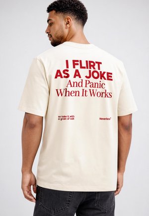 Mann mit kurzen lockigen Haaren, der ein weißes T-Shirt mit rotem Aufdruck "I FLIRT AS A JOKE And Panic When It Works" trägt, steht vor einem schlichten Hintergrund.
