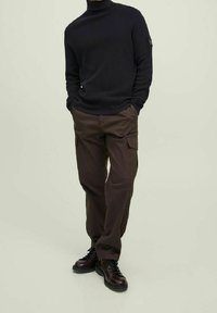 Donker geribbelde turtleneck trui gecombineerd met bruine cargo broeken met zijzakken, gedragen met donkere veterschoenen tegen een lichte achtergrond.