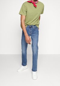 T-shirt en coton vert, jean en denim bleu, baskets blanches. T-shirt avec un petit logo, bandana noué autour du cou, coupe décontractée.